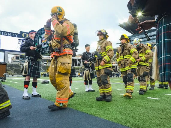Firefighters and children standing on a football field, with firefighters in full gear and children dressed in traditional Scottish attire, during a community event.