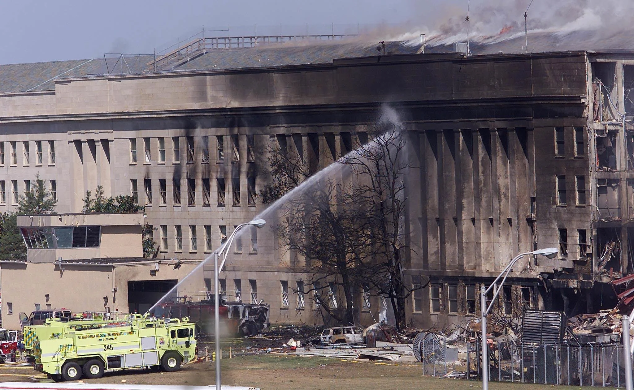 Building on fire with smoke billowing from the top. Firefighters spraying water on the flames. Emergency vehicles on the ground. Burned debris and damaged structures around the building.