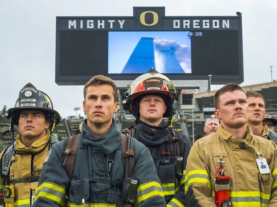 Four firefighters in uniform standing in front of a stadium scoreboard that reads "MIGHTY OREGON" with the Oregon Ducks logo.
