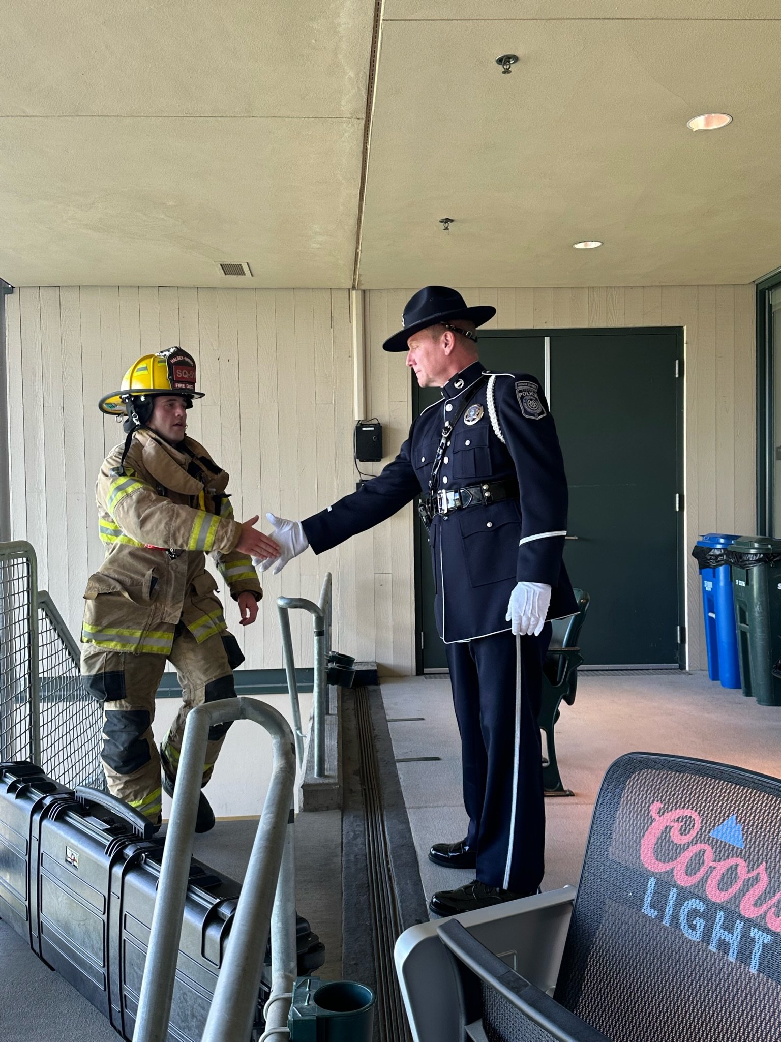 A firefighter and a police officer shake hands in an outdoor setting, with some chairs, tables, and trash bins visible around them.