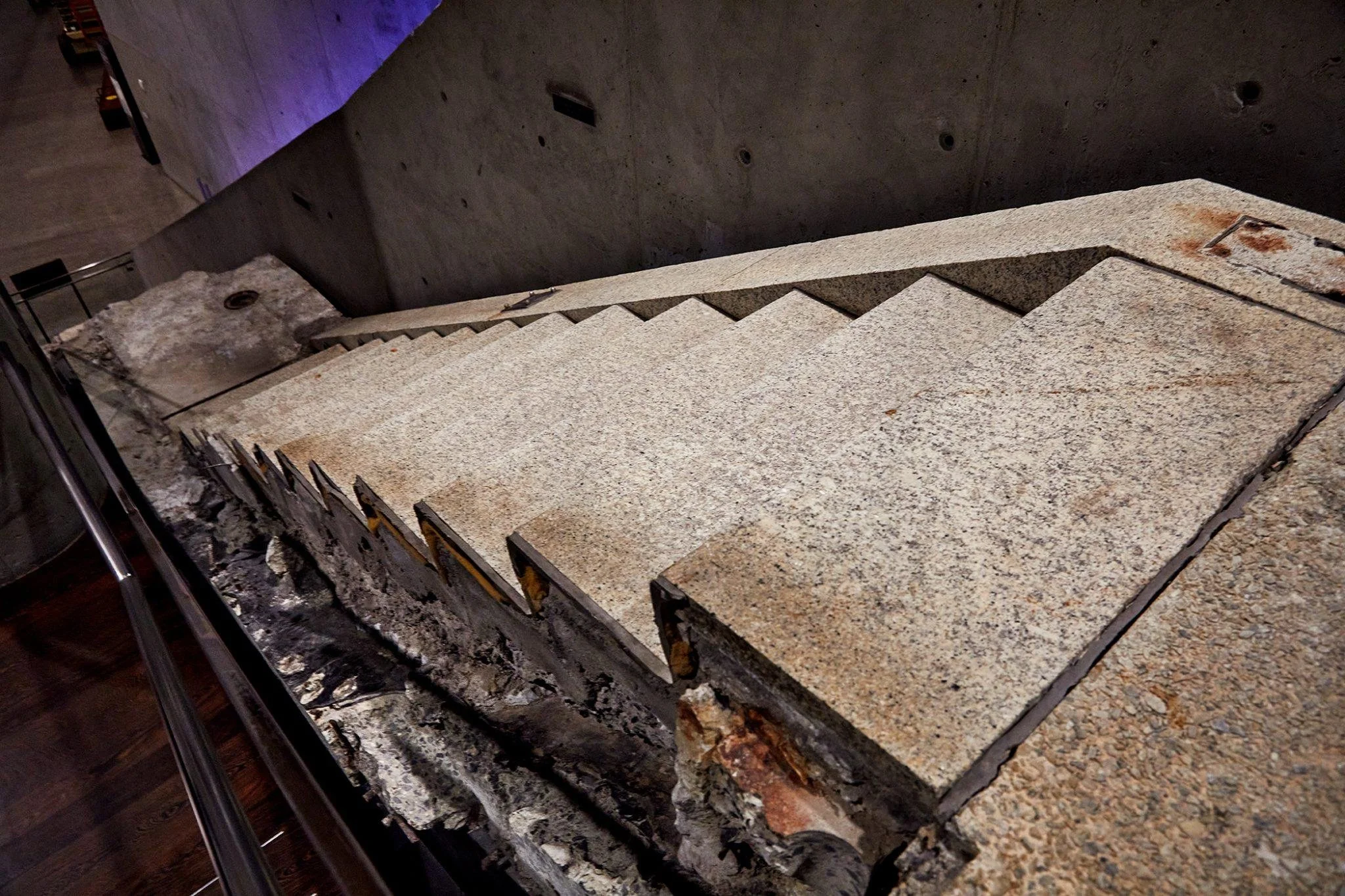 A granite staircase with a rusty metal railing, viewed from above in a dimly lit environment with concrete walls.