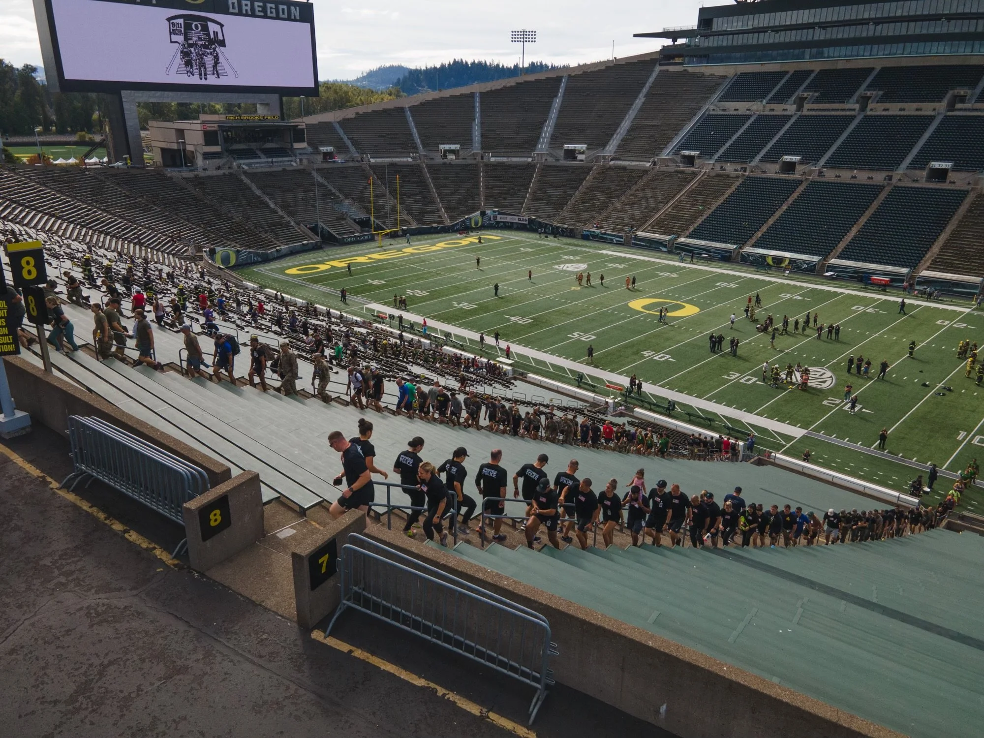 Empty Oregon Ducks football stadium with seating, field, and a large digital screen displaying an illustration of the stadium entrance. People are walking down the stairs and police officers are present.