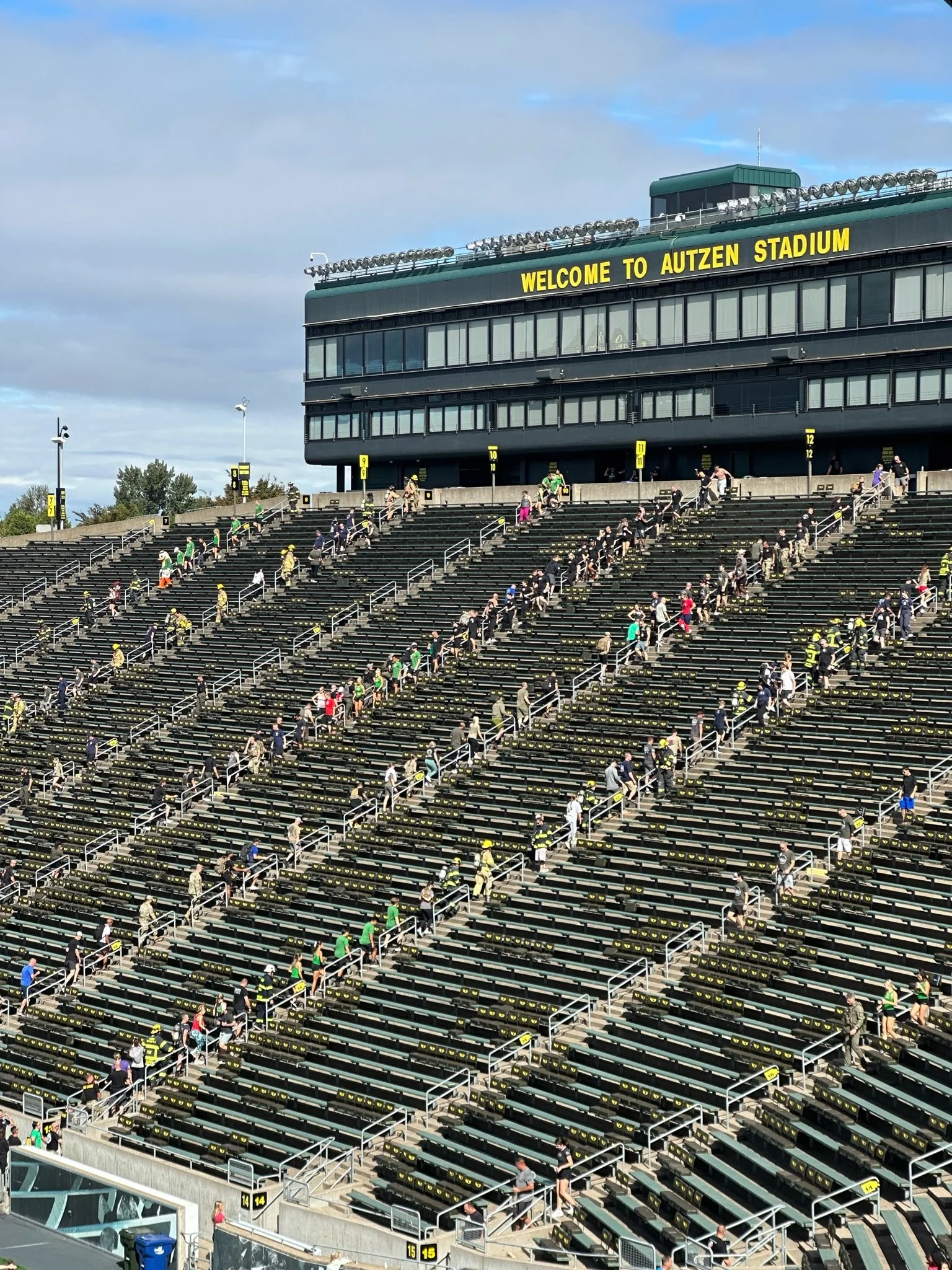 People climbing stairs at Autzen Stadium, with a building displaying a welcoming sign and the sky in the background.
