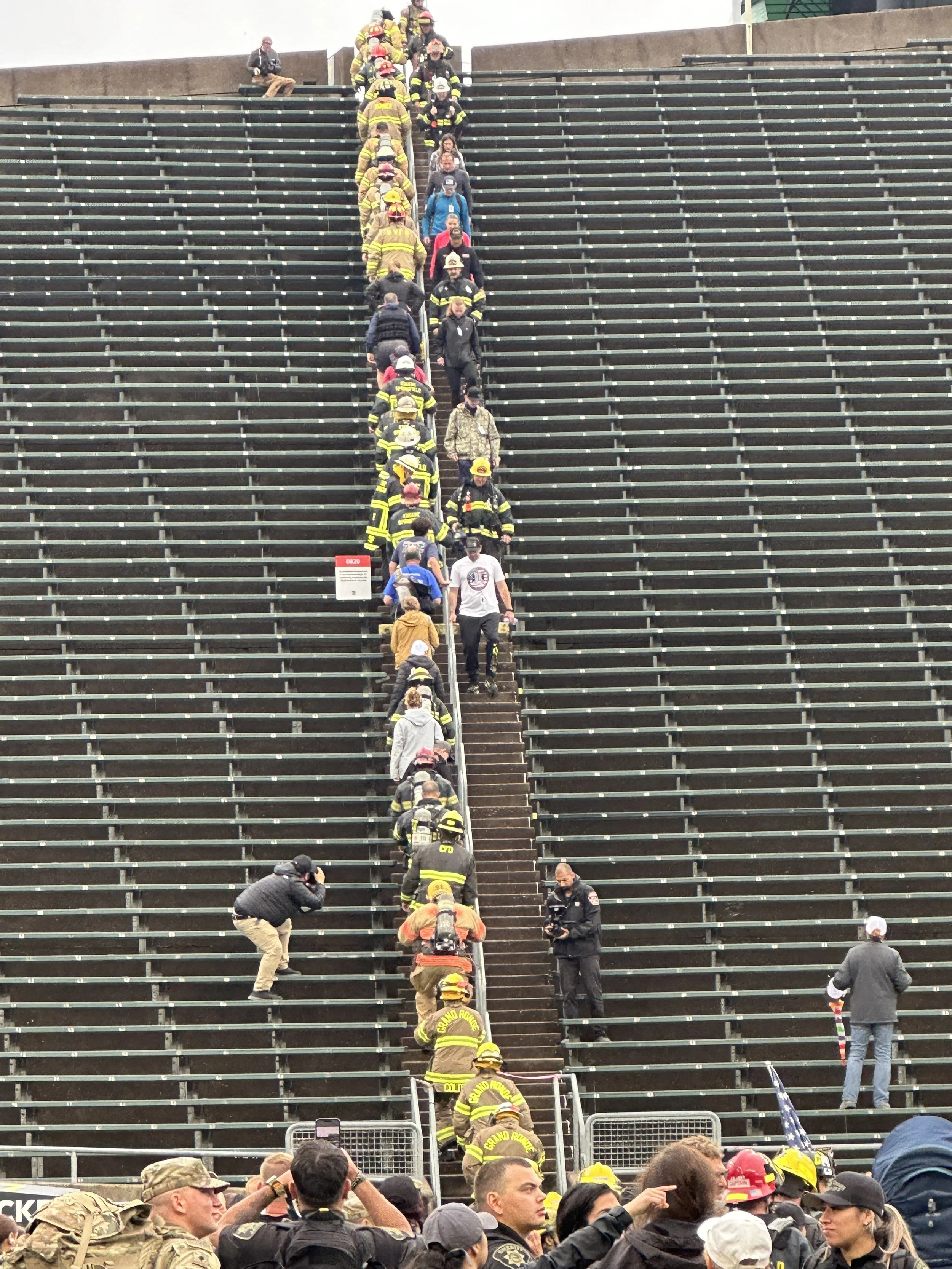 A group of firefighters and emergency responders climbing a staircase on bleachers at a public event, with spectators and photographers in the foreground.