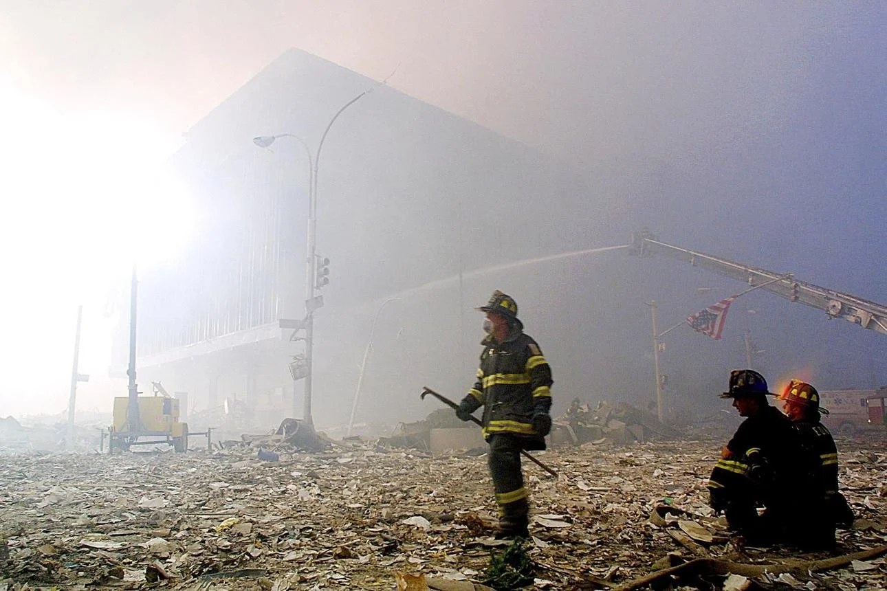 Firefighters amid debris at a disaster site with dust and smoke in the air, a damaged building, and the American flag hanging from a crane.
