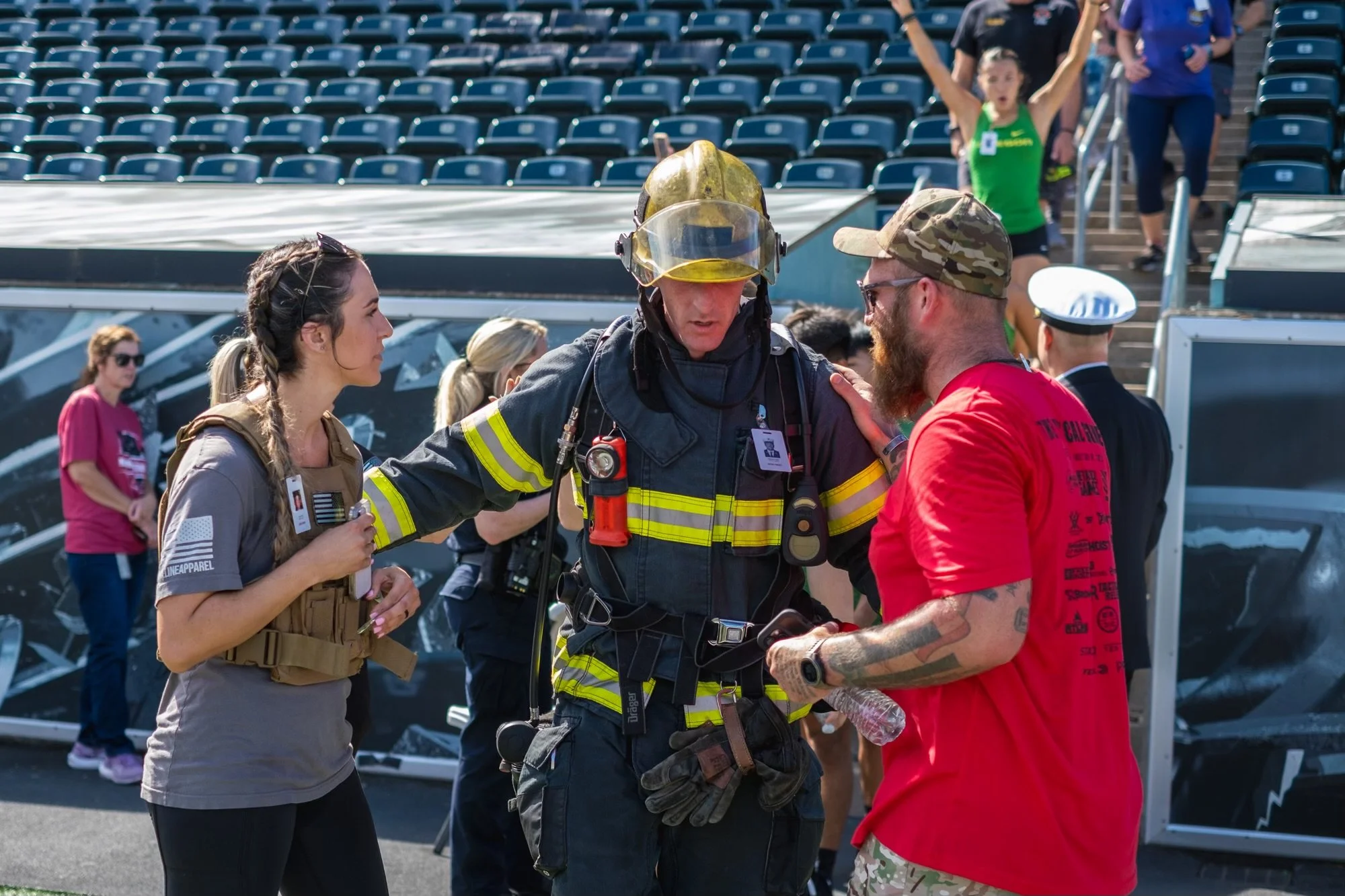 Emergency responder in firefighting gear talking to a man with tattoos in a red T-shirt at an event with empty stadium seats in the background.