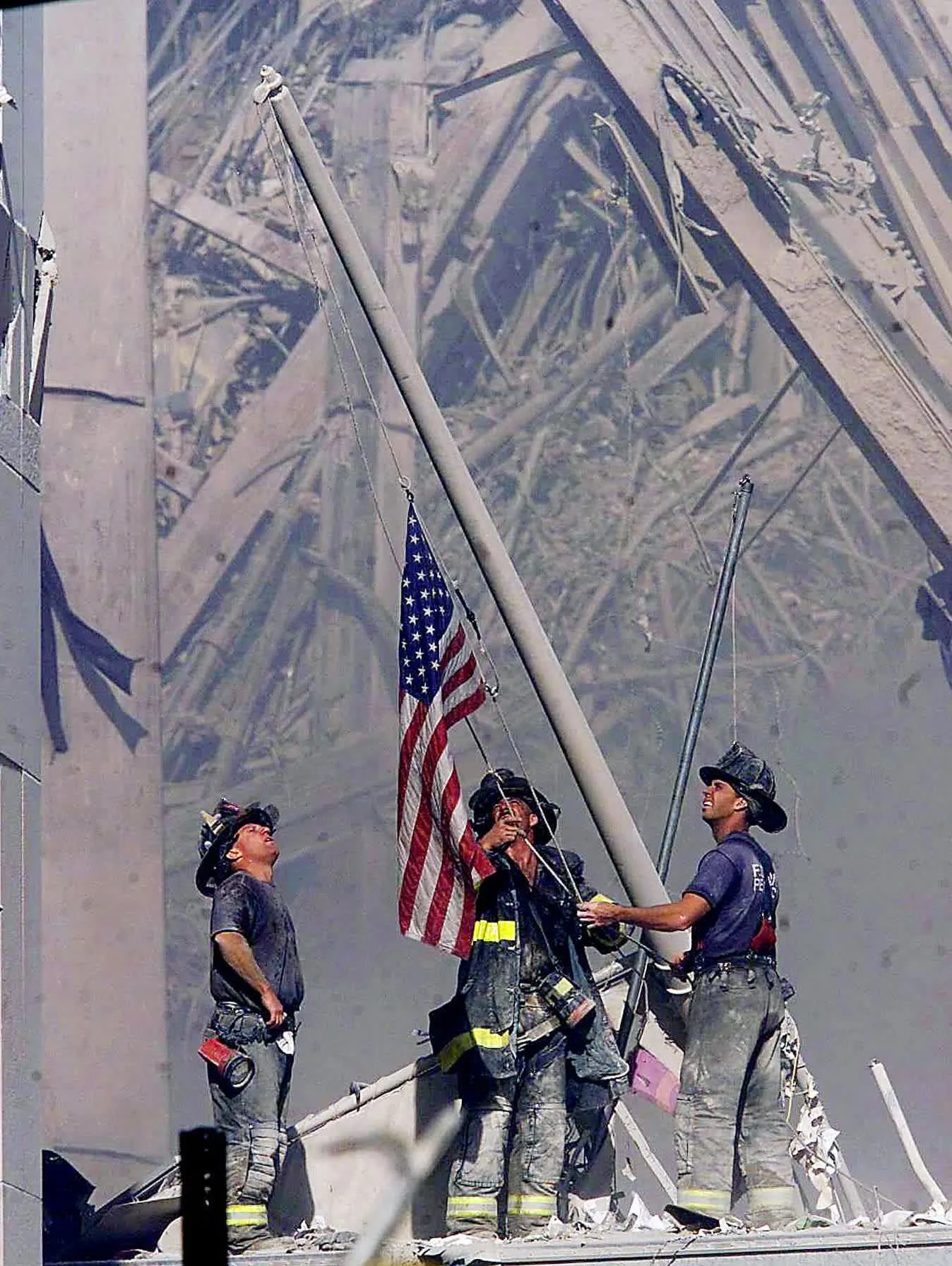 Firefighters raising the American flag at the World Trade Center site after 9/11 attacks, with a reflective surface showing the firefighters and the flag.
