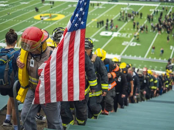 Firefighters and first responders walking up stadium stairs, with an American flag, at a football field event.