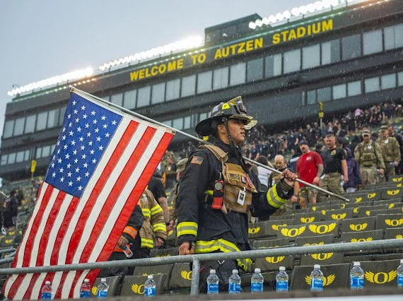 Firefighter walking with American flag in front of Autzen Stadium during daytime event with spectators.