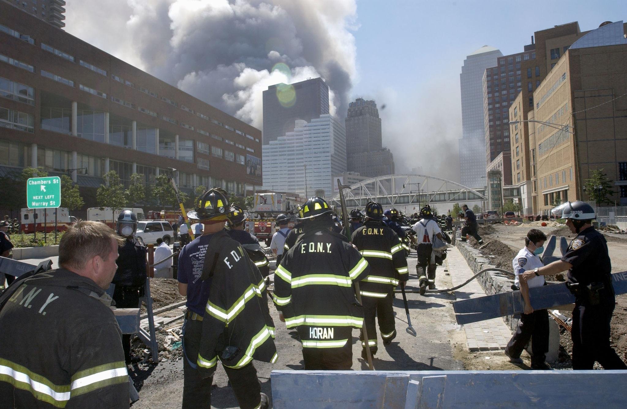 Firefighters in protective gear working on a city street affected by a fire or explosion, with smoke billowing from buildings in the background and emergency vehicles present.