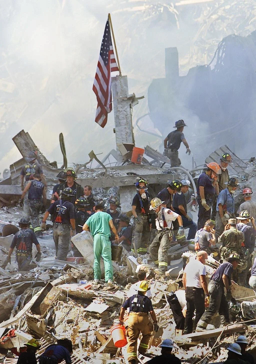 Rescue workers in safety gear search through the debris of a collapsed building, with an American flag in the background and smoke in the air.