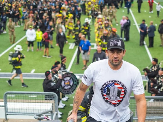 Man in white t-shirt and cap walking down stairs at a crowded outdoor event with firemen and children in the background.