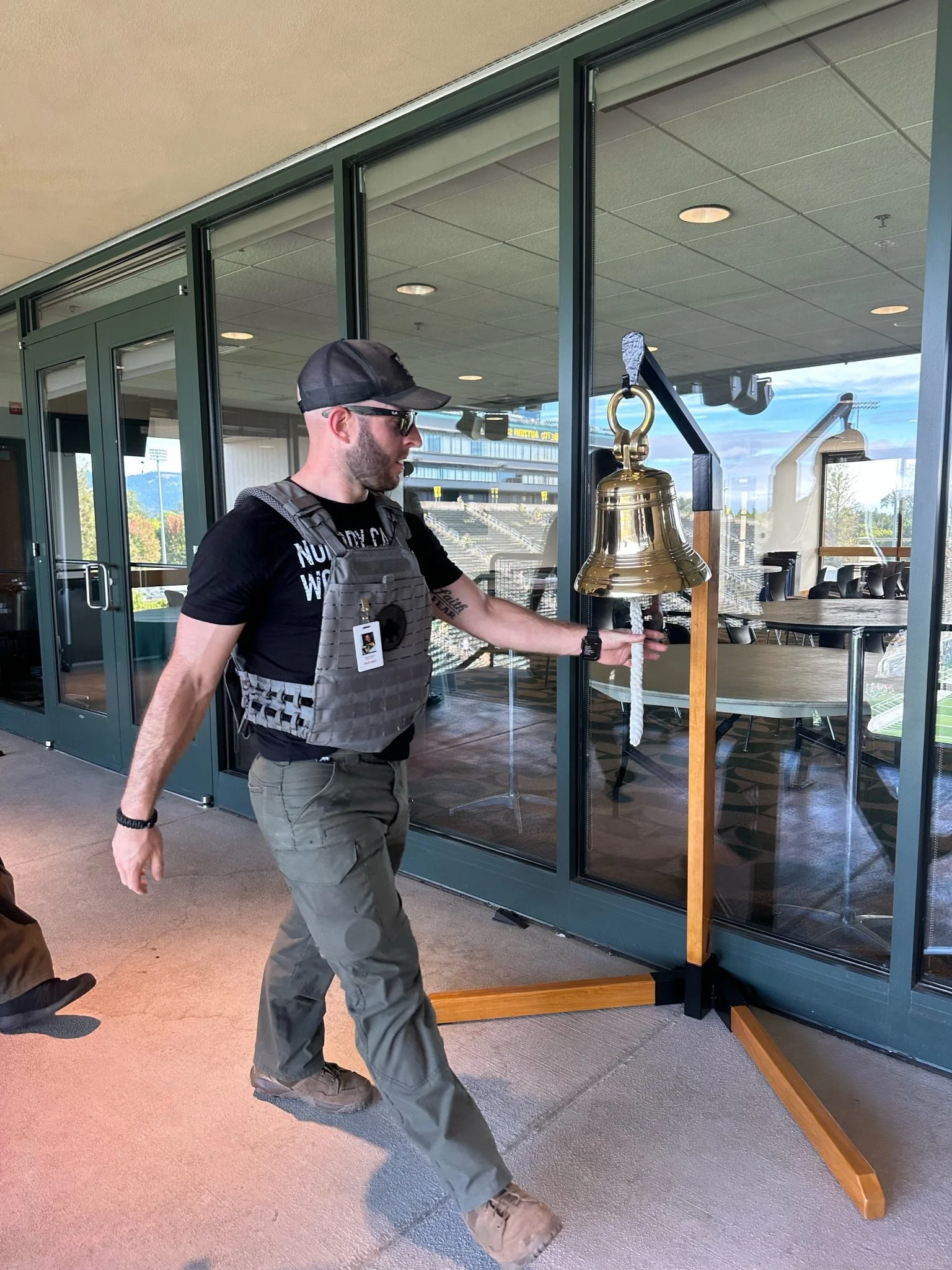 A man in tactical gear ringing a brass bell inside a glass-walled building.