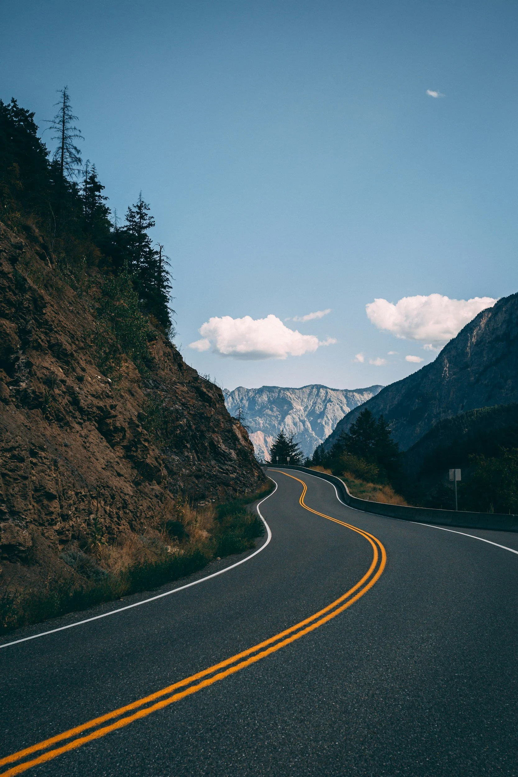 Winding mountain road with double yellow lines, bordered by rocky terrain and pine trees, under a blue sky with a small cloud.