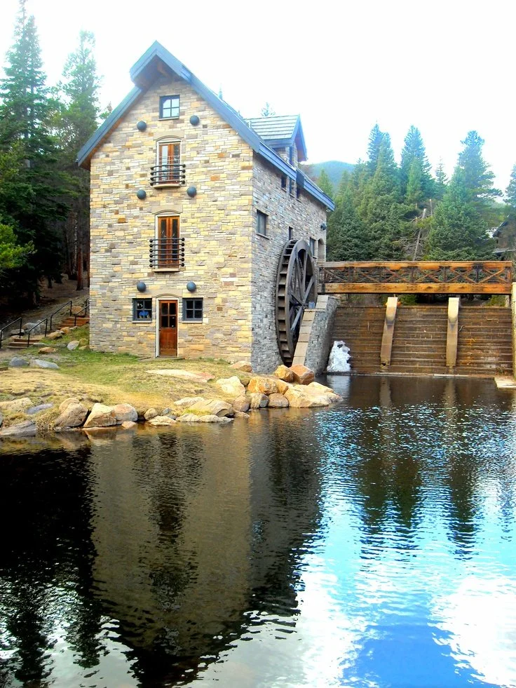 A stone watermill house beside a river, surrounded by trees with a mountainous background.