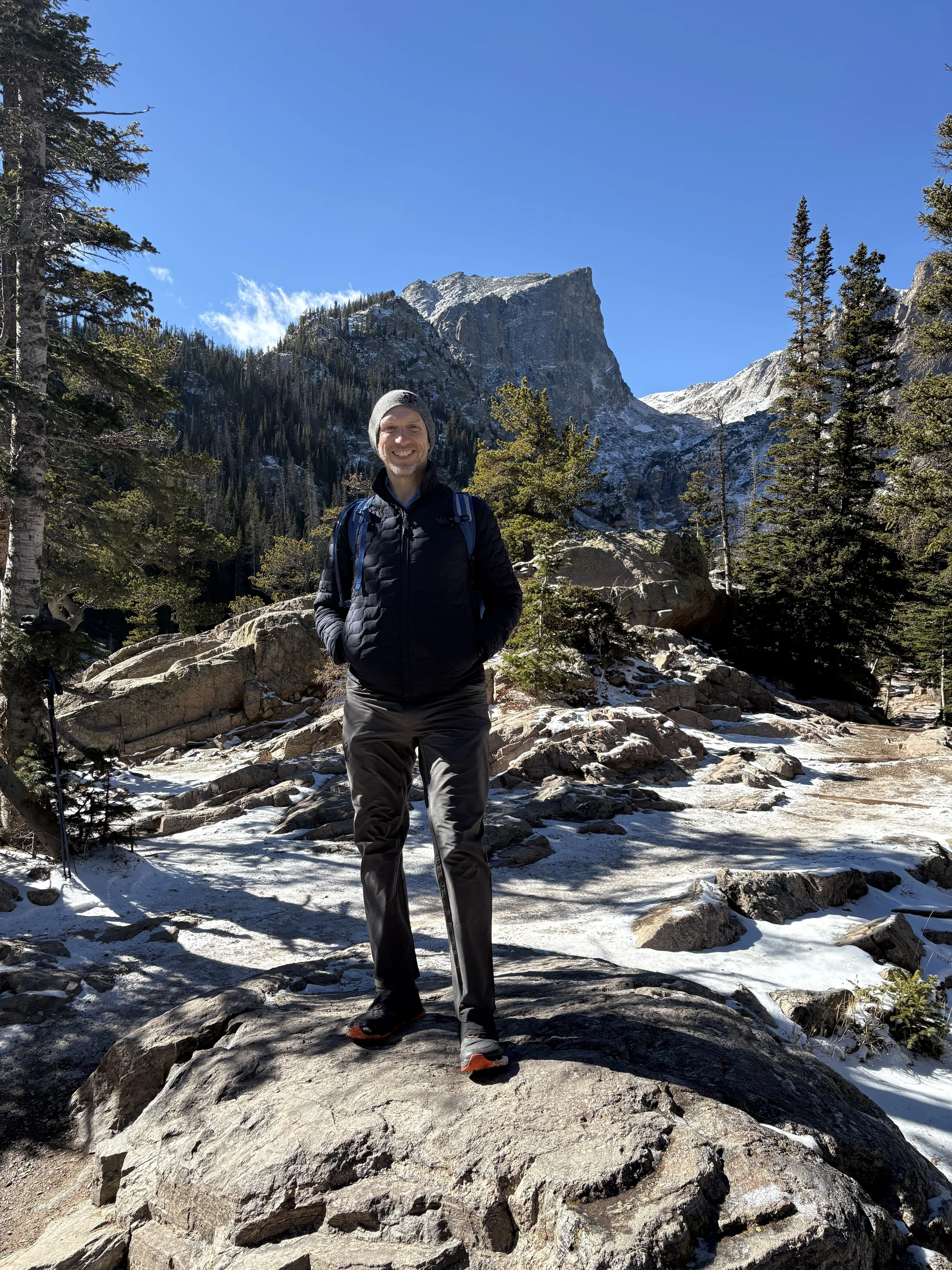 A man smiling and standing on rocky and snowy terrain in a mountainous forest landscape
