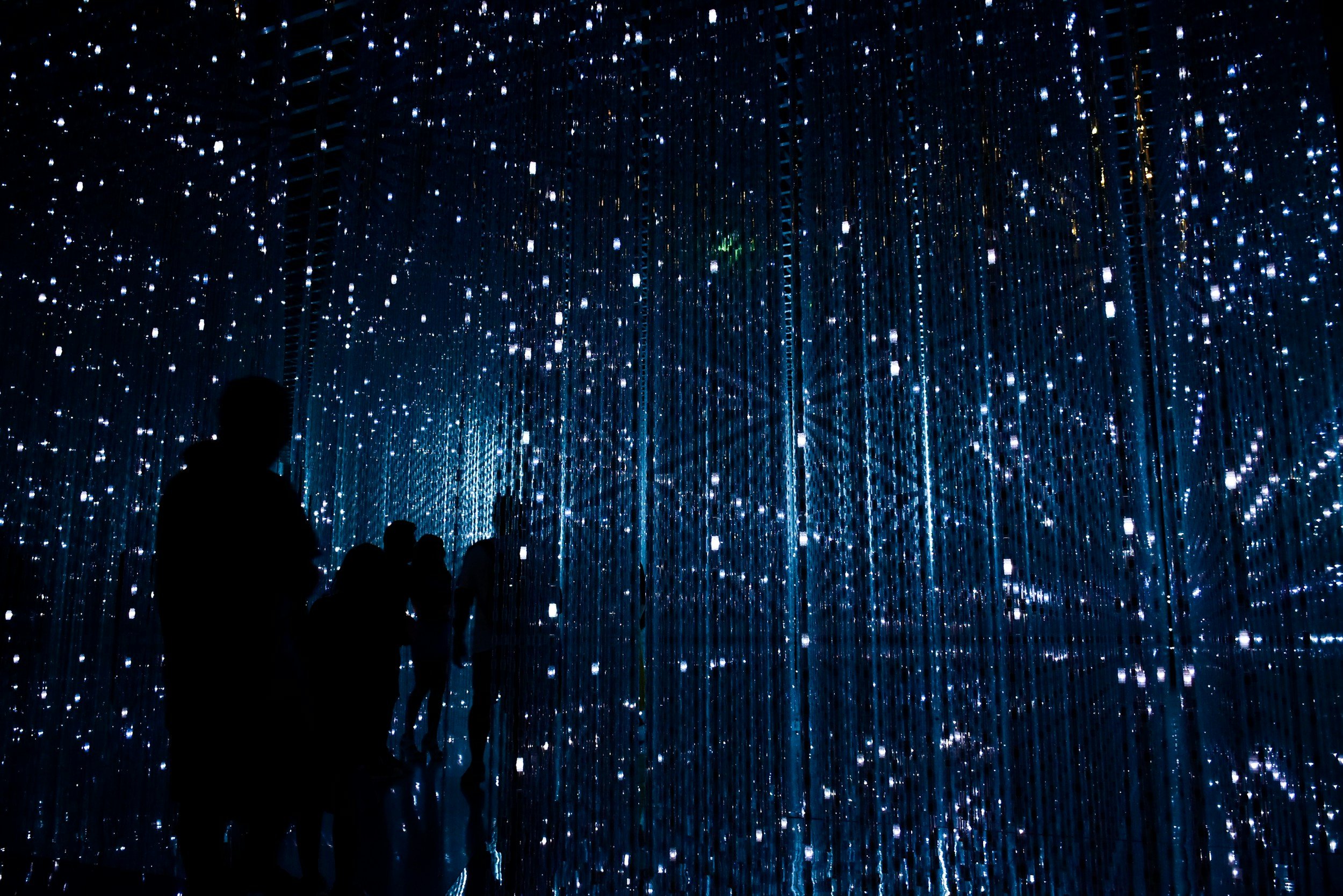Silhouetted visitors stand and walk through an immersive digital art installation filled with hundreds of glowing blue-white lights suspended in a dark mirrored room, creating the illusion of an endless starry space.