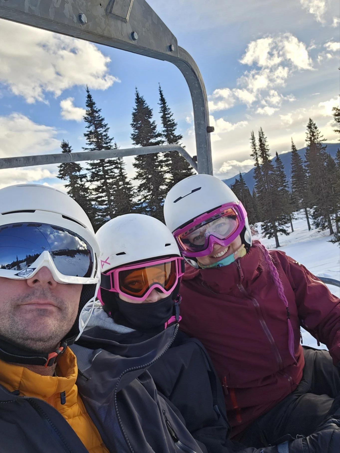 Three people wearing ski helmets and goggles, sitting on a ski lift with snow-covered mountains and pine trees in the background, under a partly cloudy sky.