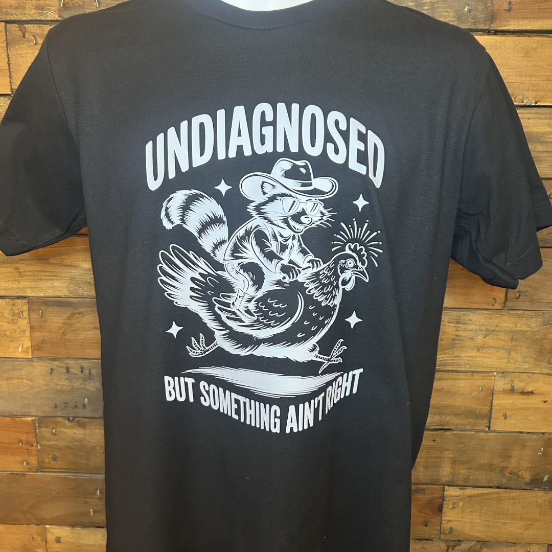 Black T-shirt featuring a graphic of a raccoon with sunglasses and a hat, riding on top of a chicken. The text above the graphic reads 'UNDIGNOSED' and below it says 'BUT SOMETHING AIN'T RIGHT'.