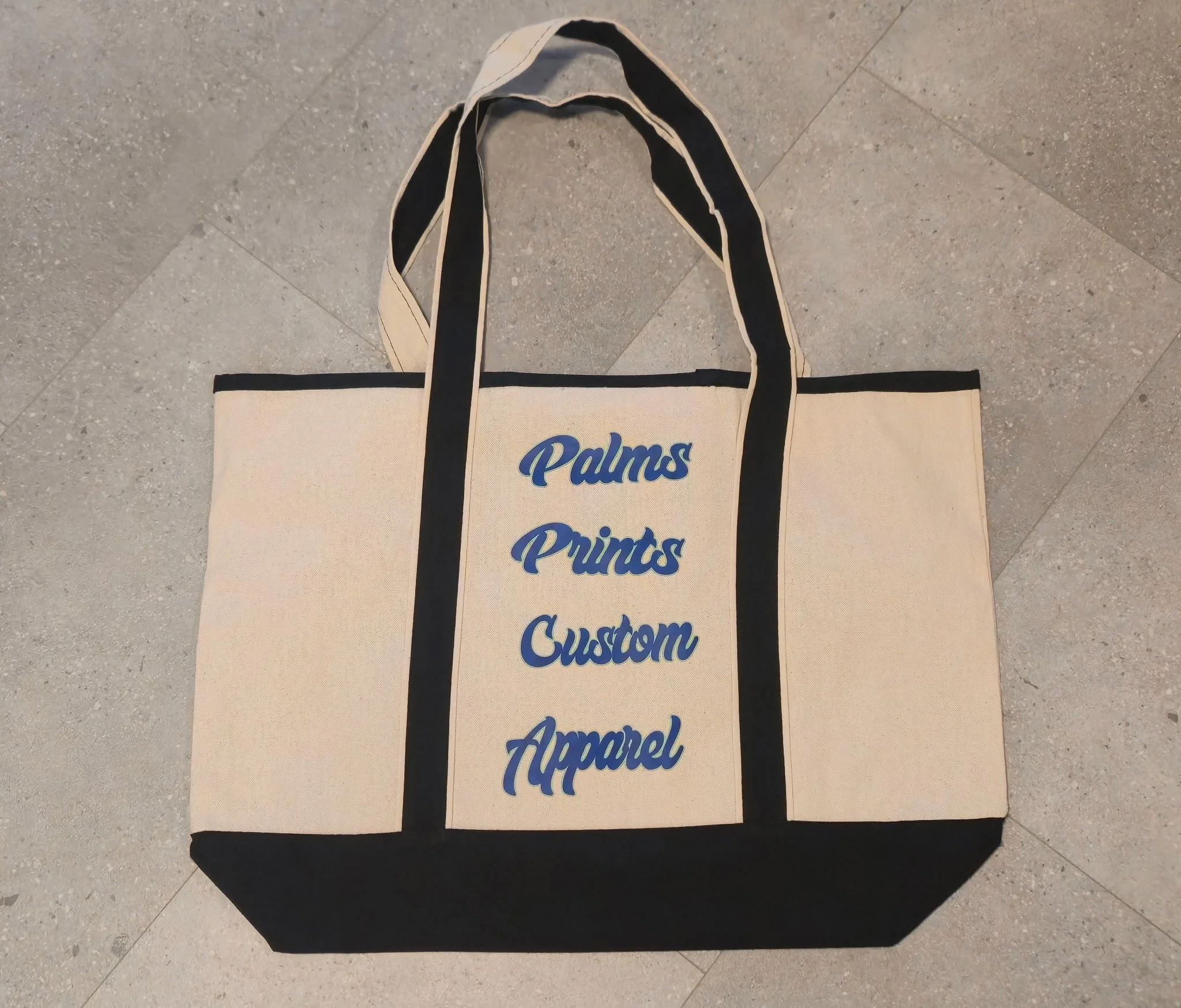 A beige canvas tote bag with black handles lying on a tiled floor. The bag has the words "Palms" and "Prints" written in blue cursive font.