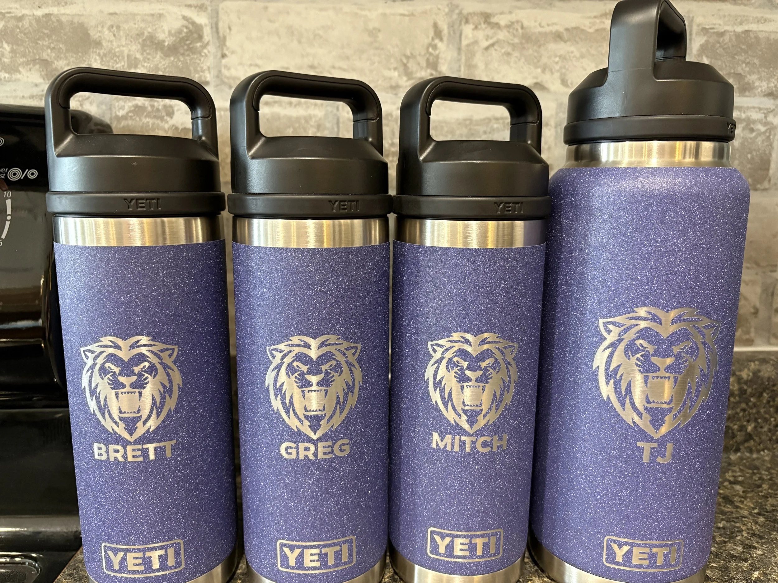 Four personalized Yeti water bottles with lion logos and names 'Brett,' 'Greg,' 'Mitch,' and 'TJ' printed below the logos, arranged in a row on a shelf.