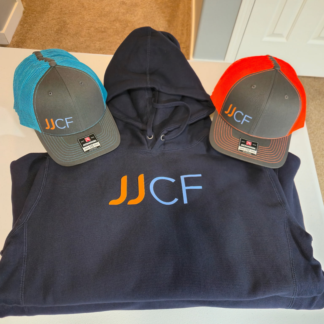 A hoodie and two baseball caps with the letters JJCF, one in orange and blue and the other in orange and white, placed on a white surface.