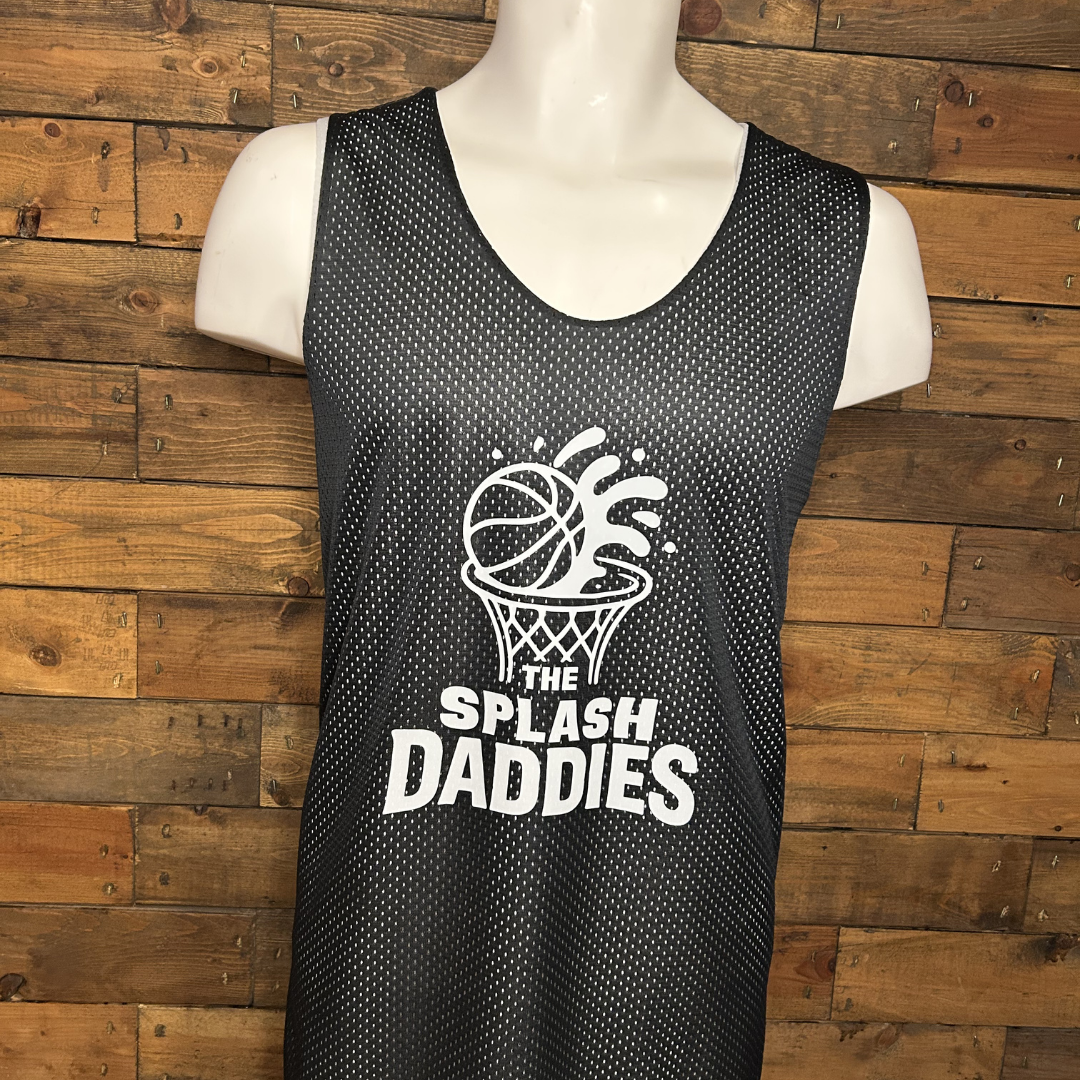 Black sleeveless basketball jersey with white text and logo that says 'The Splash Daddies,' featuring a basketball going into a hoop with splash effects, displayed on a mannequin against a wooden wall.