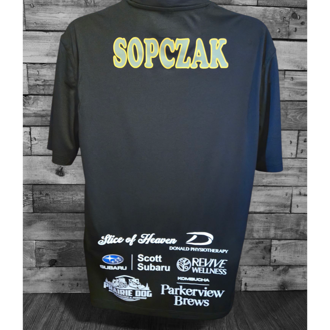 Back of a black sports jersey with the name 'SOPCZAK' in yellow and white text at the top. Various sponsor logos and brands are displayed at the bottom, including Subaru, Scott Subaru, Prairie Dog, Revive Wellness, Parkerview Brews, and others, against a wooden wall background.