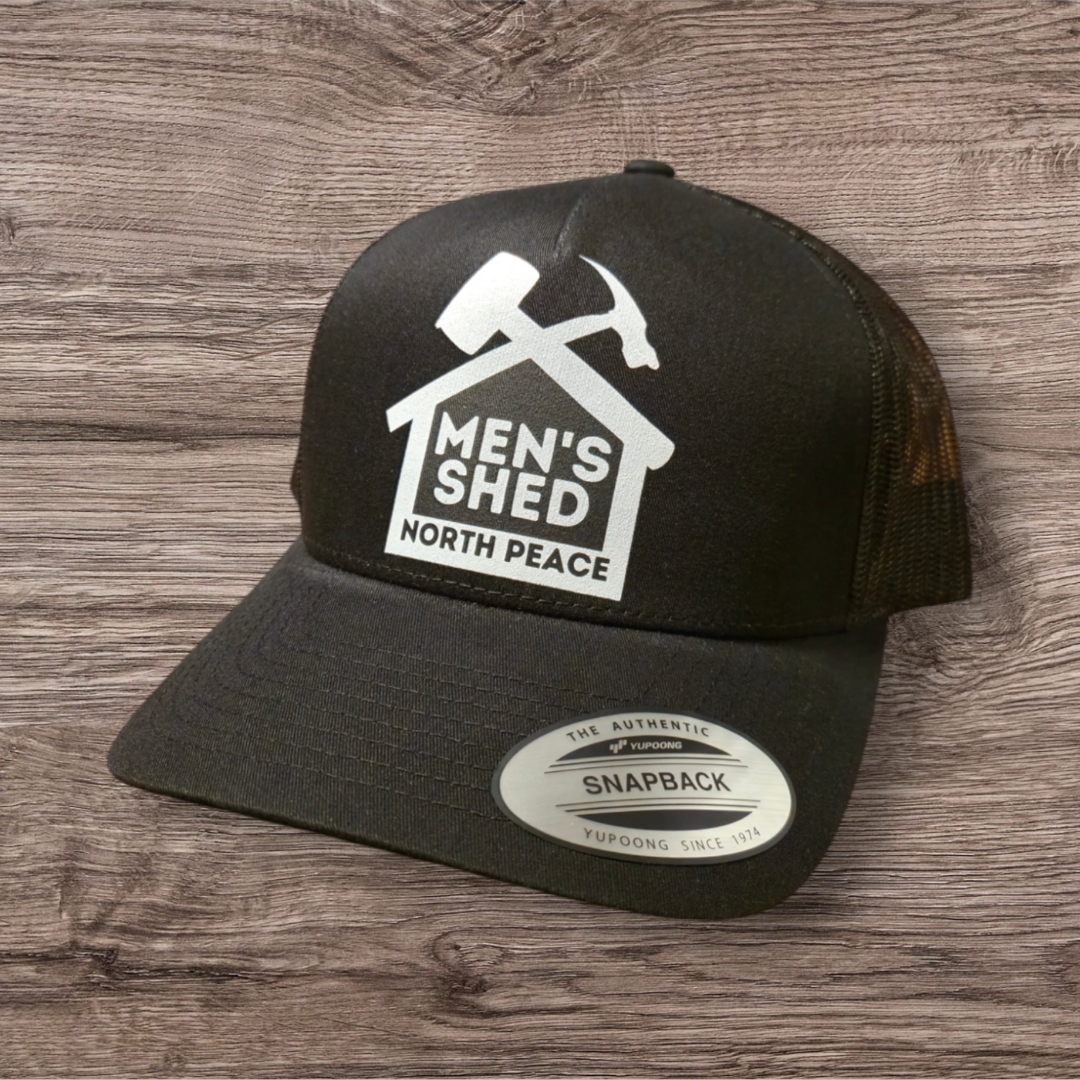 Black snapback cap with a patch that reads 'Men's Shed North Peace' and features a hammer, screwdriver, and house icon on a wooden surface.
