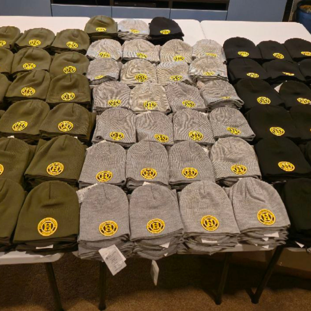 Multiple knit beanies with yellow emblems, stacked on a table in green, gray, and black colors.