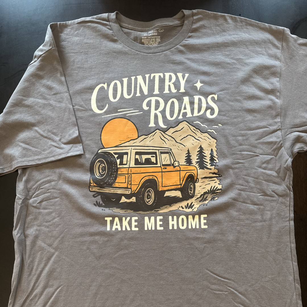 Grey T-shirt with a design featuring an orange off-road vehicle, mountains, pine trees, a setting sun, stars, and text 'Country Roads, Take Me Home.'