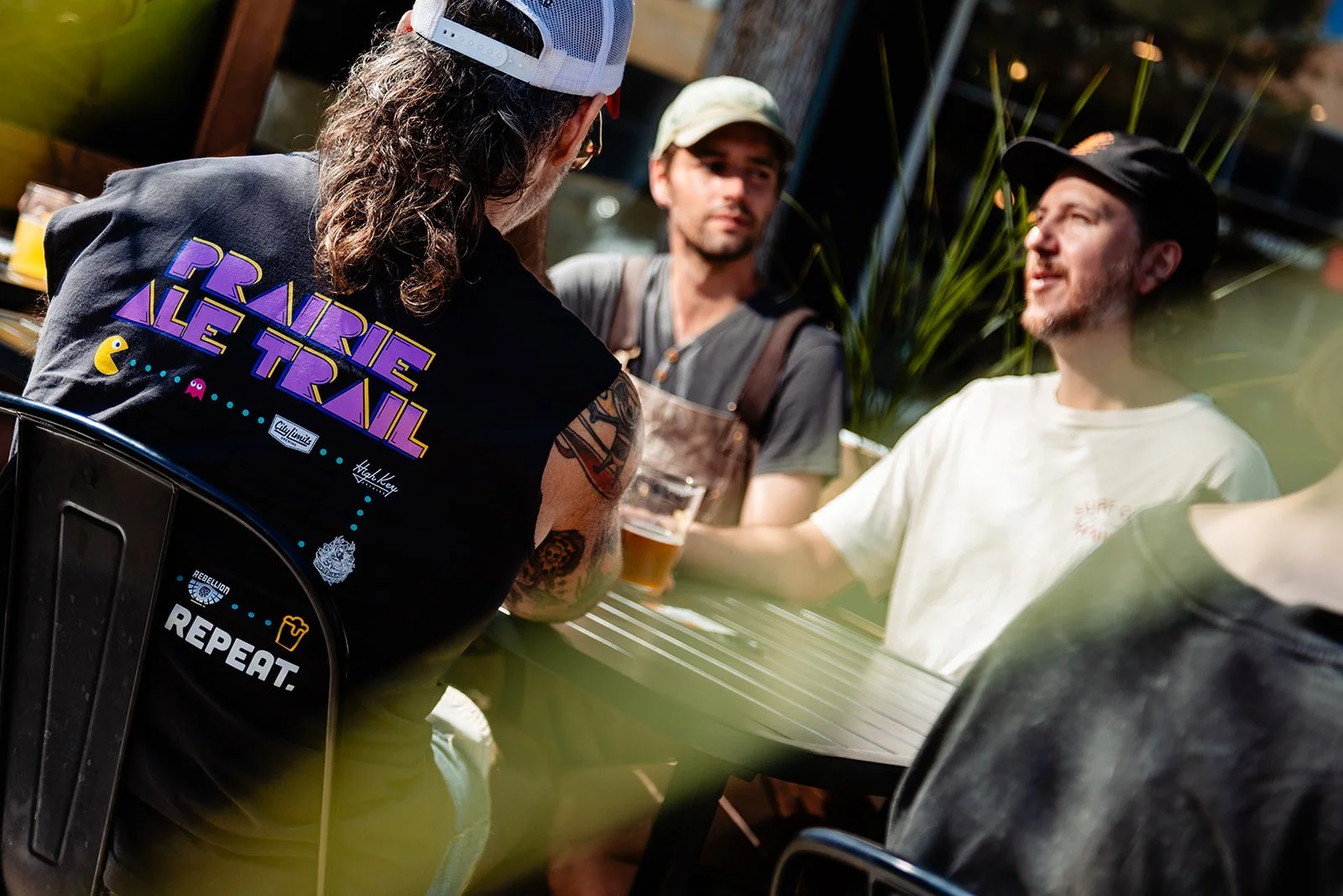 Four men are sitting outdoors at a table, engaging in conversation, with one holding a glass of beer. They are casually dressed, with some wearing caps, and the setting appears to be a bar or outdoor cafe.