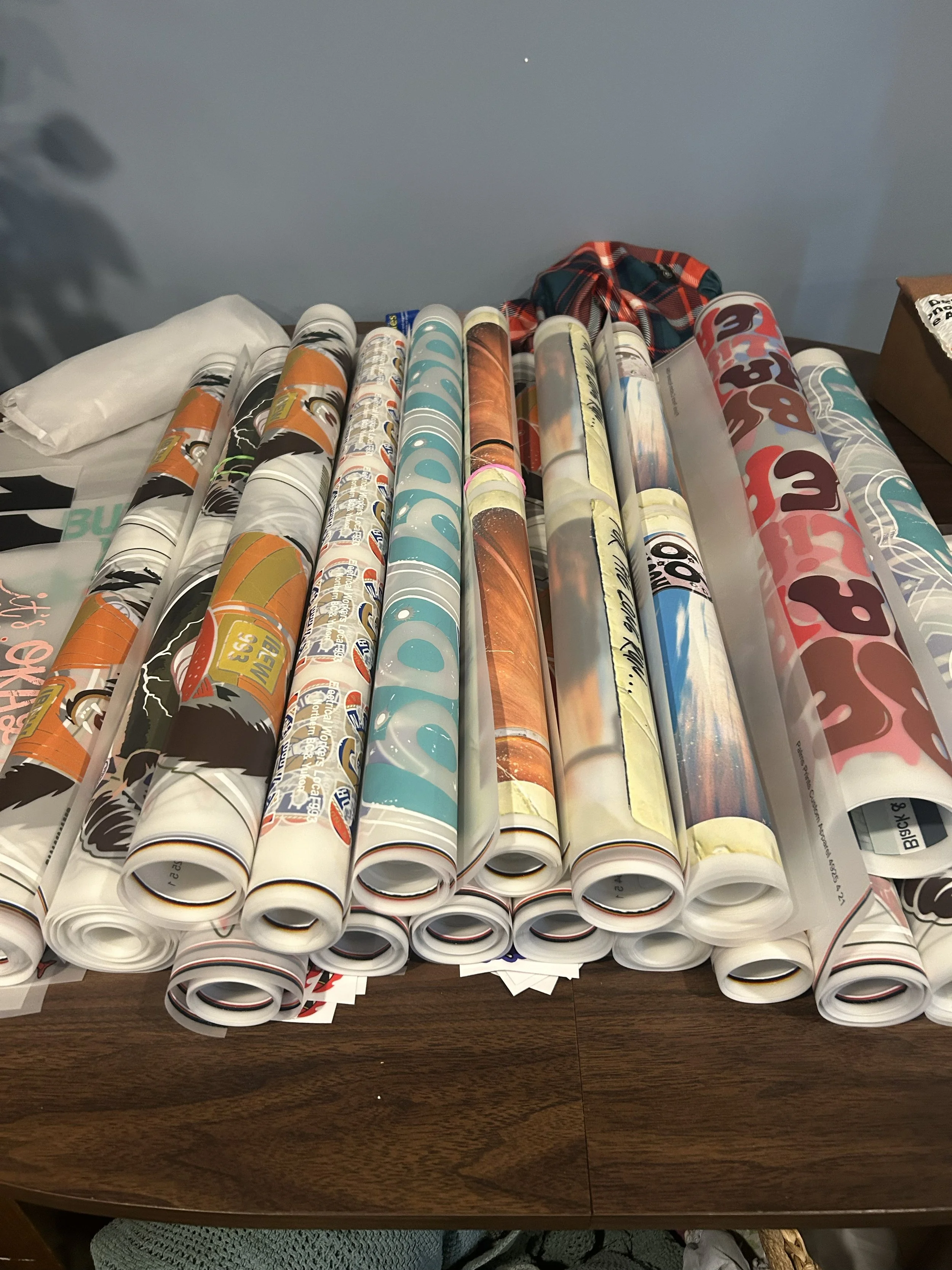 A table covered with multiple roll of stickers or decals with various designs and colors, some with orange, blue, brown, white, pink, and gray shades. A plaid cloth is partially visible behind the rolls. The background features a plain wall with a small bright spot.