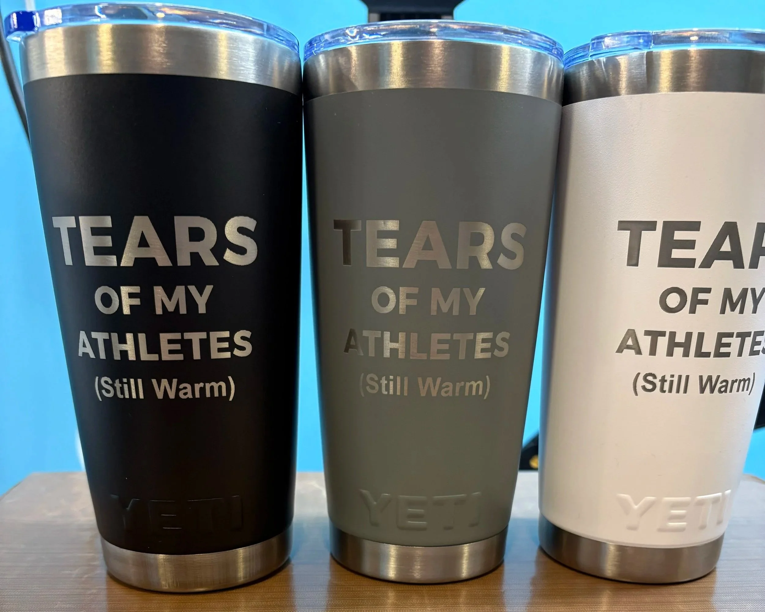Three travel mugs with stainless steel bottoms and colored bodies, than have the words "TEARS OF MY ATHLETES" written on them, with the words on the black mug in white, the middle mug in gold, and the right mug in white, all on a dark surface against a light blue background.