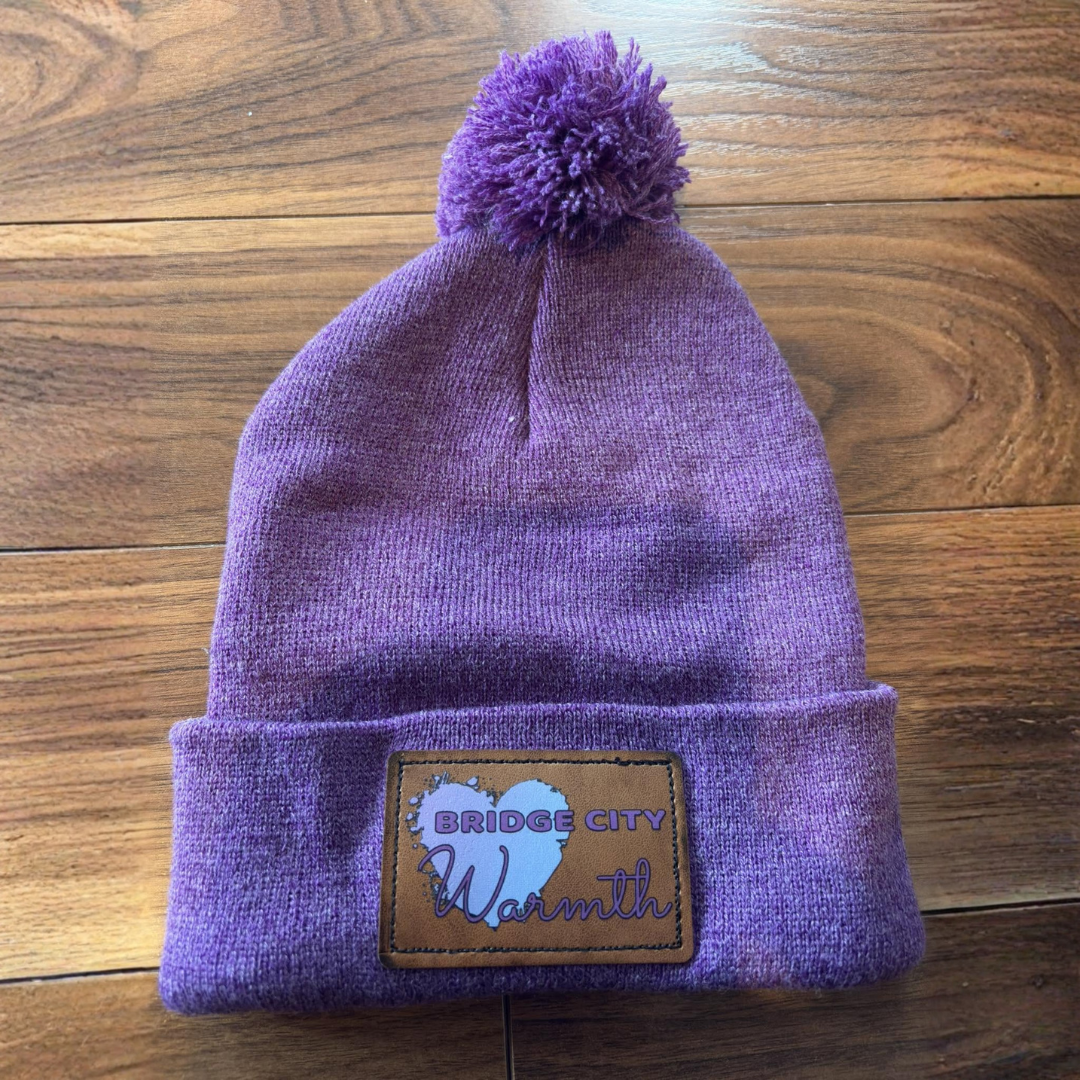 Purple knit beanie with a pom-pom on top, featuring a brown patch reading "Bridge City Warmth" on a wooden surface.