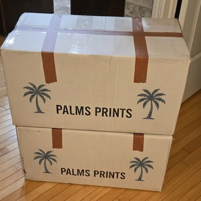 Two cardboard boxes stacked on top of each other, labeled 'PALMS PRINTS' with a palm tree graphic on each, placed on a hardwood floor in a room.