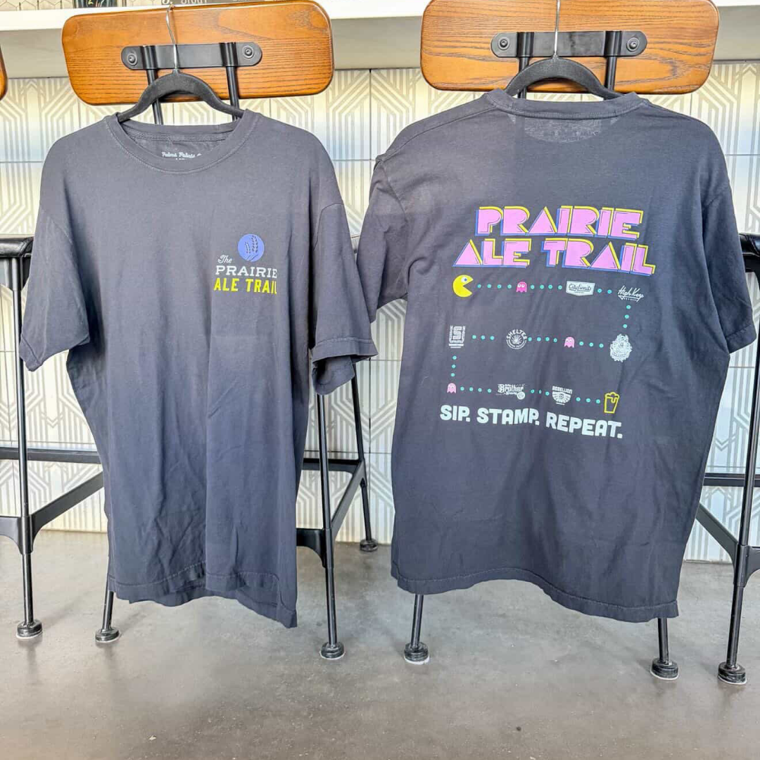 Two gray T-shirts hanging on wooden hangers on a display rack. The front of the left shirt features a small design with the text 'The PRAIRIE ALE TRAIL' and a blue circle. The back of the right shirt has colorful text 'PRAIRIE ALE TRAIL' styled like a video game, along with a Pac-Man graphic and icons of various breweries, ending with the phrase 'SIP. STAMP. REPEAT.'