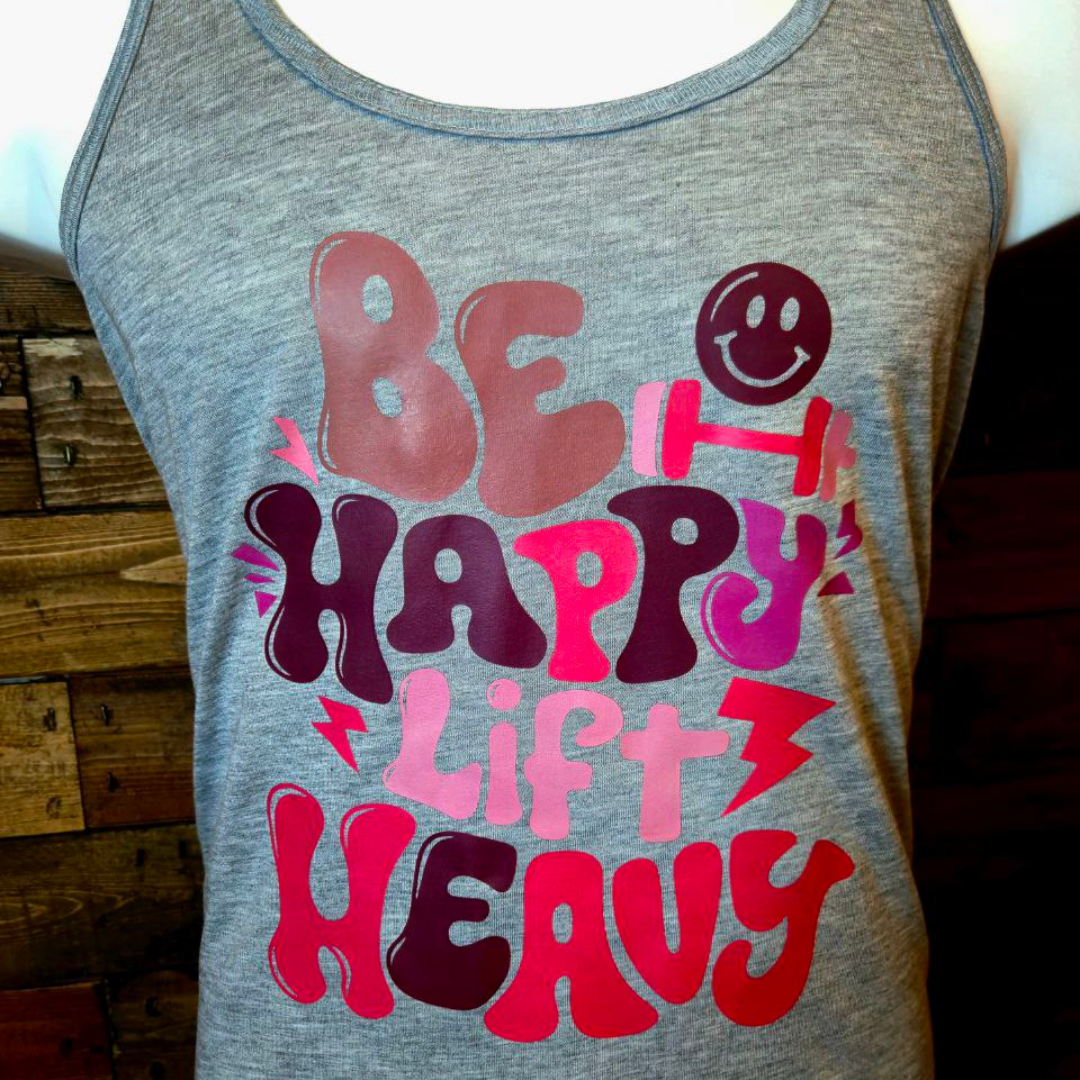 Gray tank top with colorful, playful text that reads 'Be Happy, Lift Heavy' with smiley faces and lightning bolt graphics.