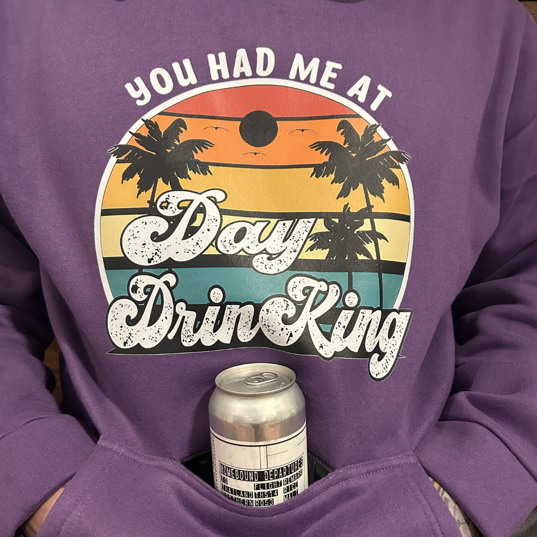 A person wearing a purple sweatshirt with a graphic that says "You had me at Day Drinkin'" featuring a sunset and palm trees. The person has a can of beverage in their pocket.