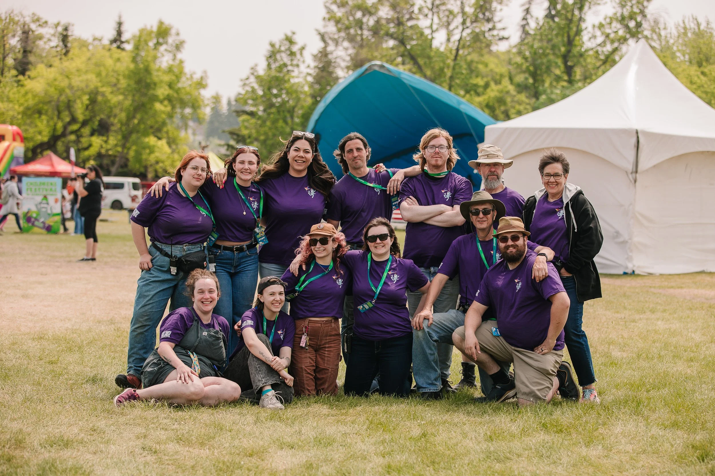 Group of people standing outdoors on grass at a festival or event, many wearing purple shirts with logo, some wearing hats and sunglasses, smiling and posing for a photo, tents and trees in the background.
