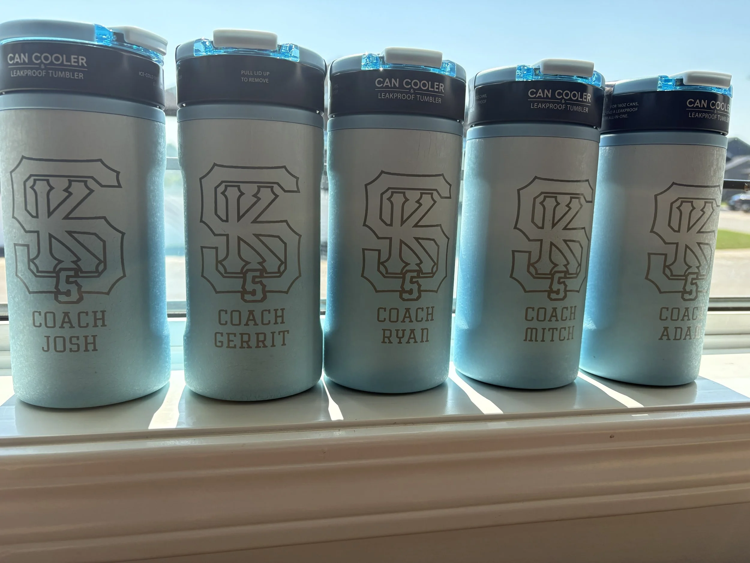 Set of white insulated tumblers with black and blue lids, labeled with names of football coaches, lined up on a windowsill with sunlight shining through.