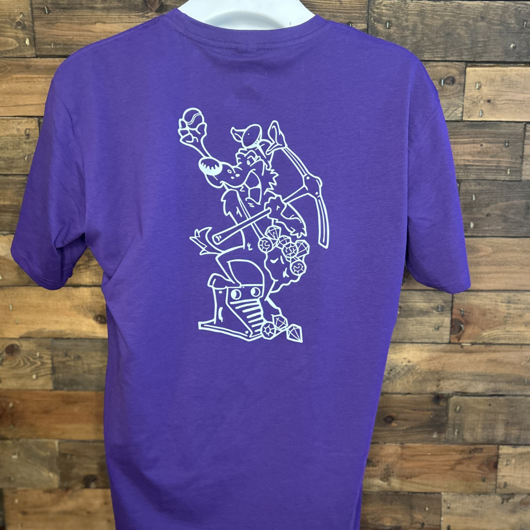 Purple T-shirt with a white line art illustration of a cartoon wolf holding a hammer and a ball, surrounded by flowers, a treasure chest, and a diamond, hanging on a wooden wall.