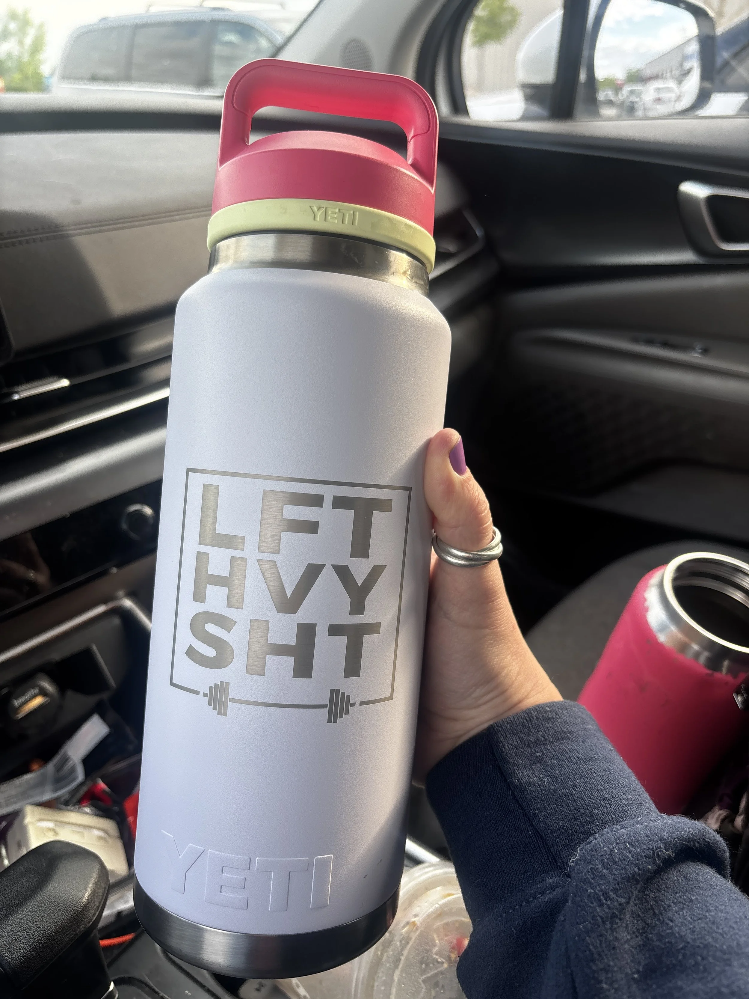 A person holding a white Yeti water bottle in a car, with a black lid. The label on the bottle has large text that reads "LIFT HEAVY SHIT" and a small dumbbell graphic. Other water bottles are visible in the car.