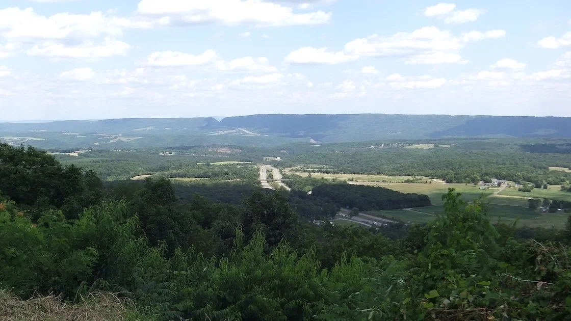 A wide landscape view with rolling green hills, a highway, and scattered trees, under a partly cloudy sky.