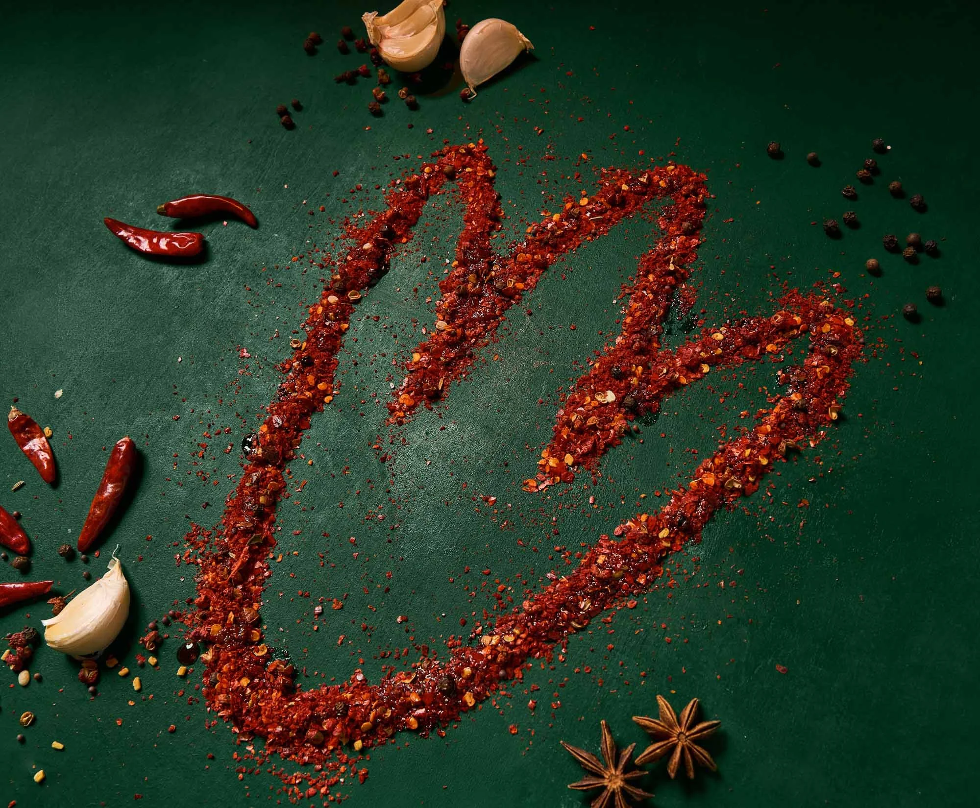 Assorted red chili peppers, garlic cloves, black peppercorns, star anise, and crushed red pepper flakes arranged on a green surface.