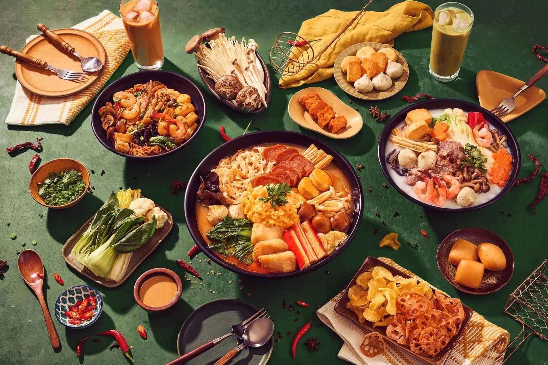 Variety of Asian dishes on a green table, including soups, noodles, vegetables, and fried snacks, with drinks and utensils.
