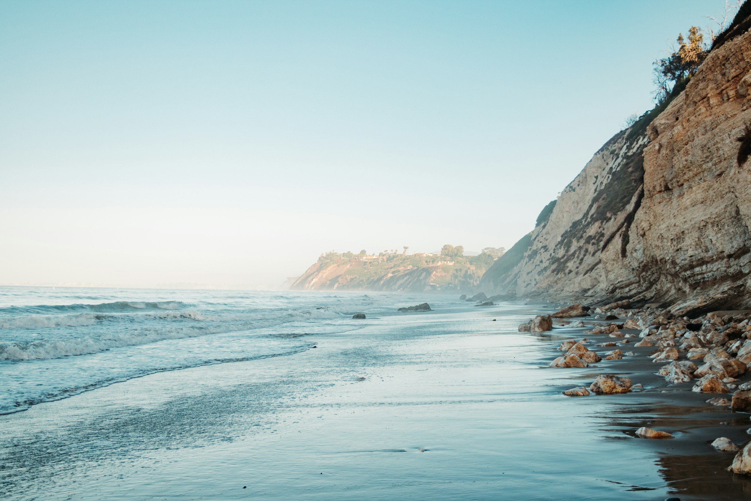 Coastal view with ocean waves, rocky shoreline, and cliffs in the distance under a clear sky.