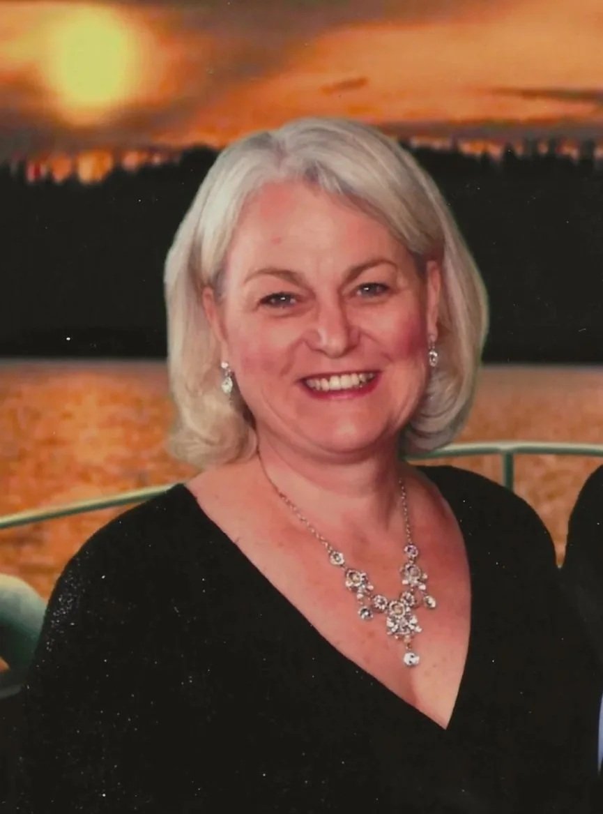 A woman with gray hair smiling in front of a sunset or sunrise over a body of water.