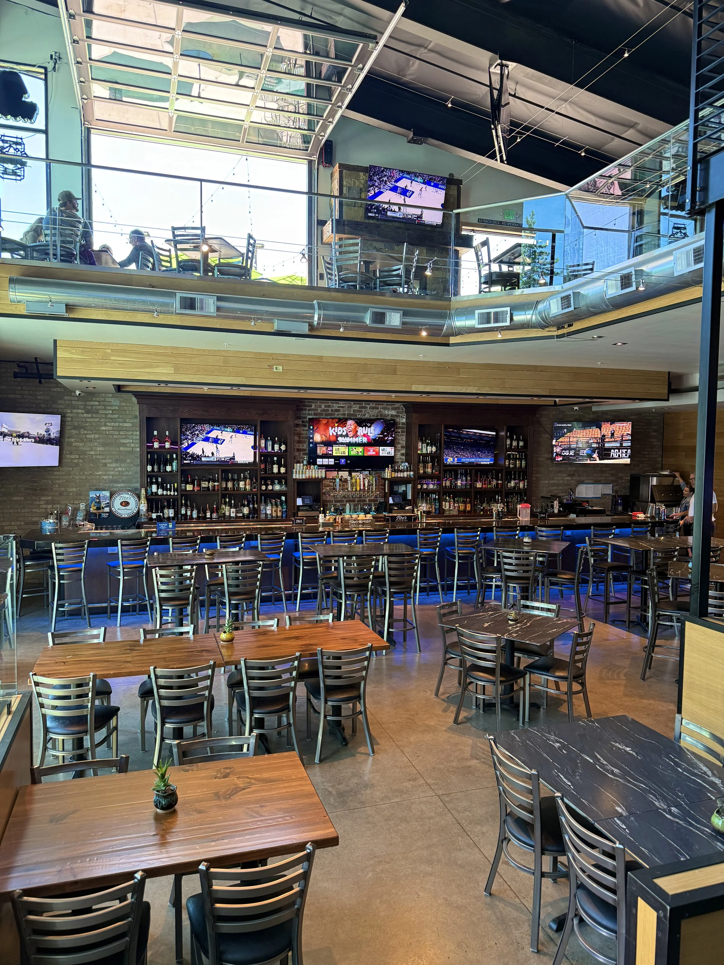 modern restaurant with tiered seating area, multiple flat-screen TVs displaying sports, and wooden tables with small potted plants, featuring environmental ductwork and large windows letting in natural light.