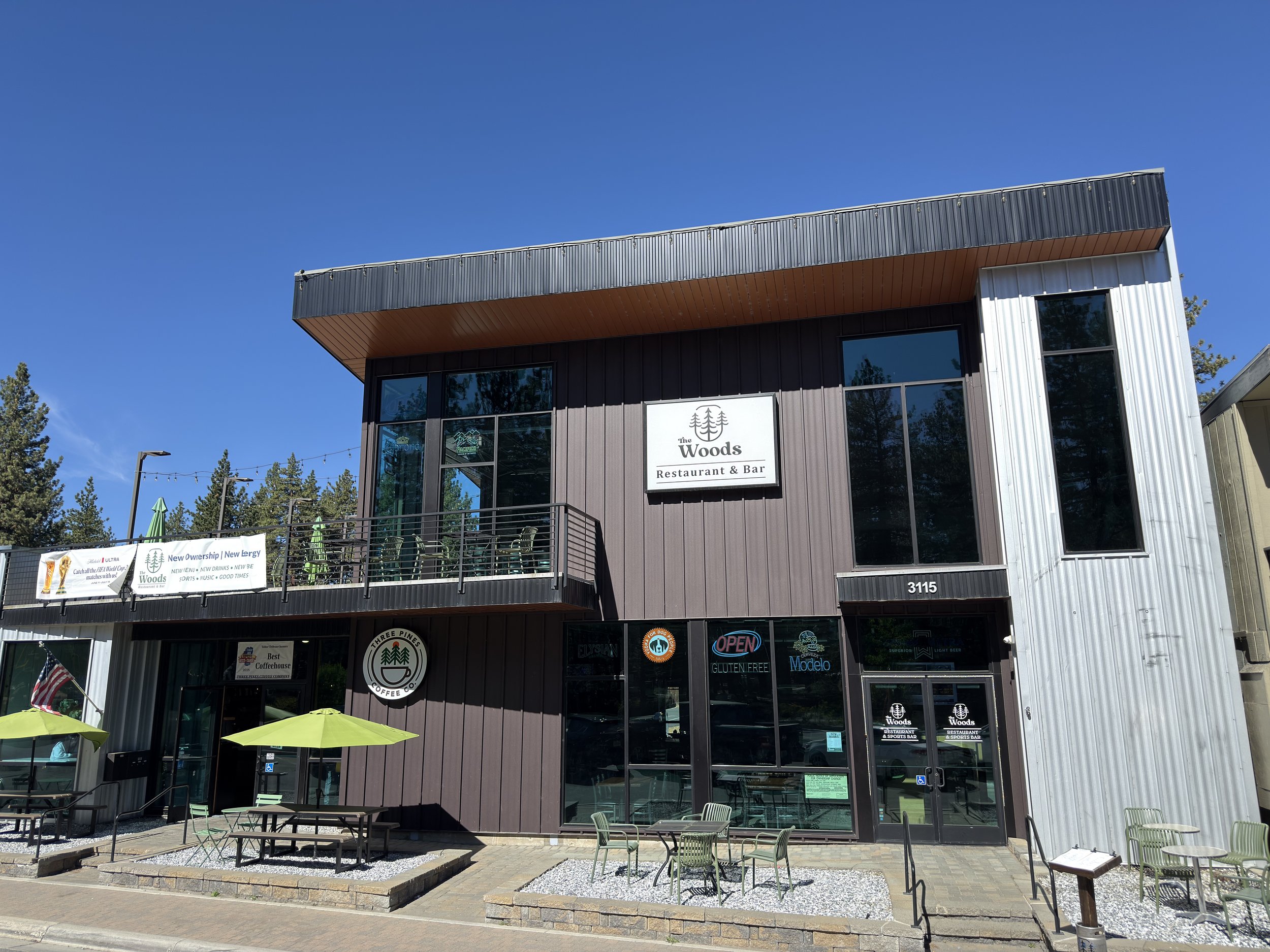 A multi-story building with a restaurant and sports bar called "The Woods," featuring large windows, outdoor seating with yellow umbrellas, and a sign indicating gluten-free options, located at 3115. The building has two distinct exterior finishes, one dark and one light metal siding, and is situated in a sunny area with trees and a clear blue sky and string lights overhead.